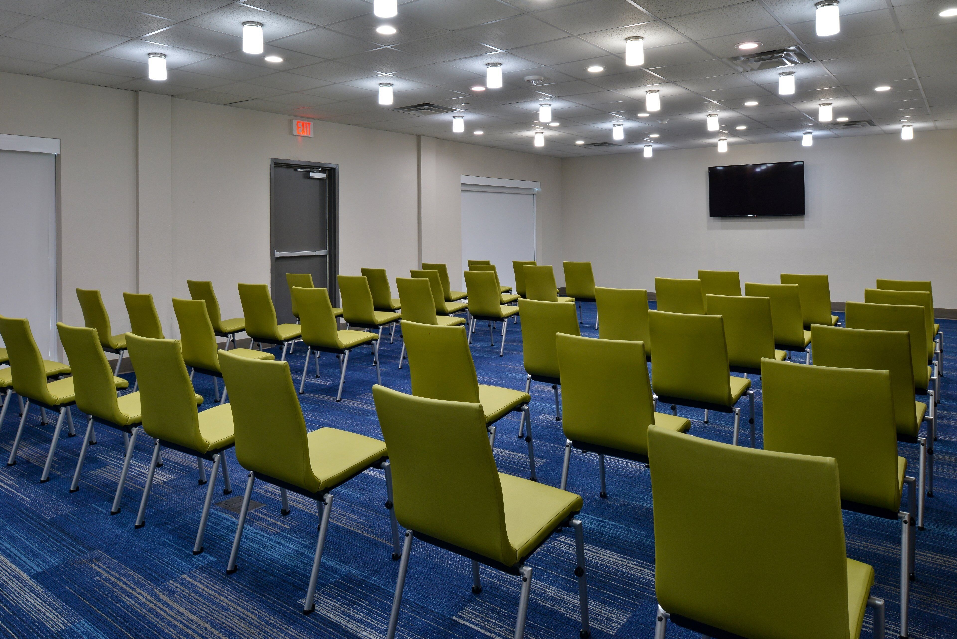 A conference room with rows of green chairs, a whiteboard, a projector screen, and a flat-screen TV on the wall.