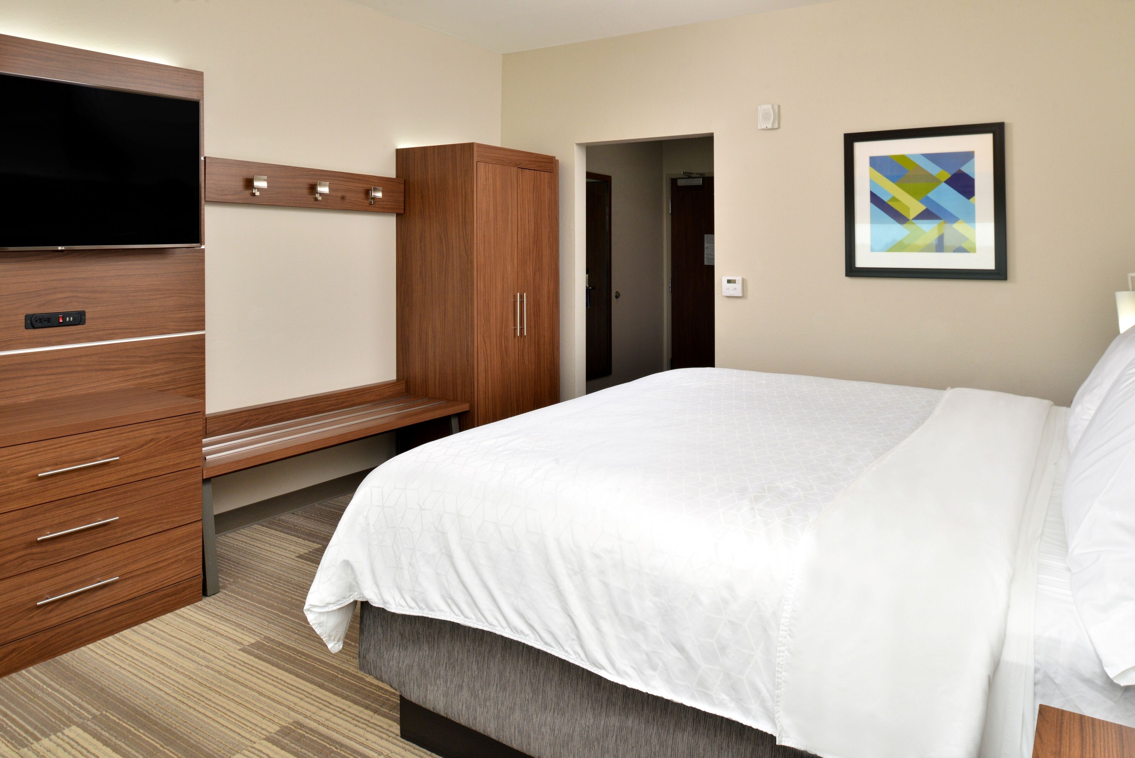 A hotel room with a bed, a wooden wardrobe, a flat-screen TV, and a framed abstract painting on the wall.