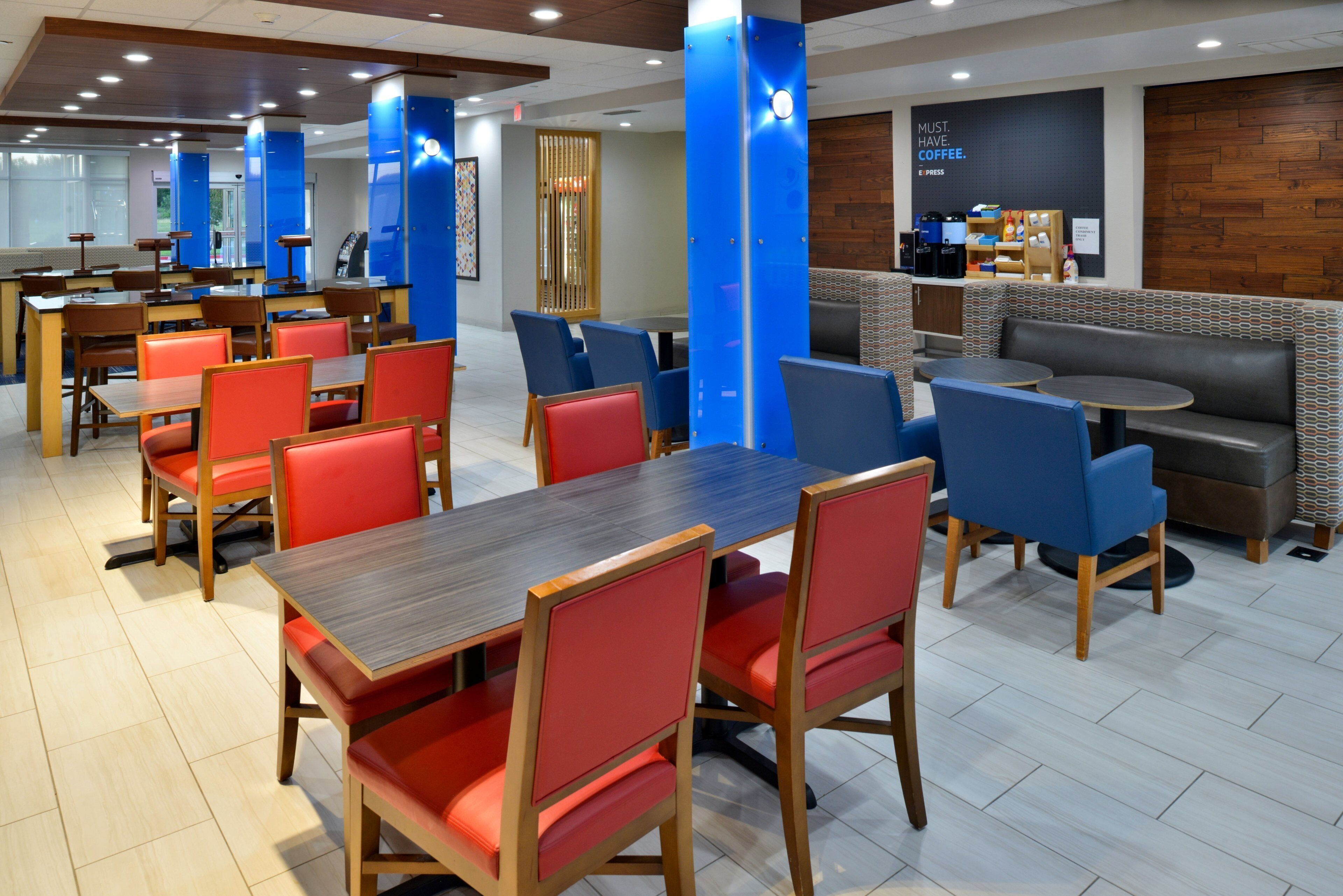 A modern restaurant with wooden tables and red and blue chairs, a coffee bar, and a seating area with patterned cushions.