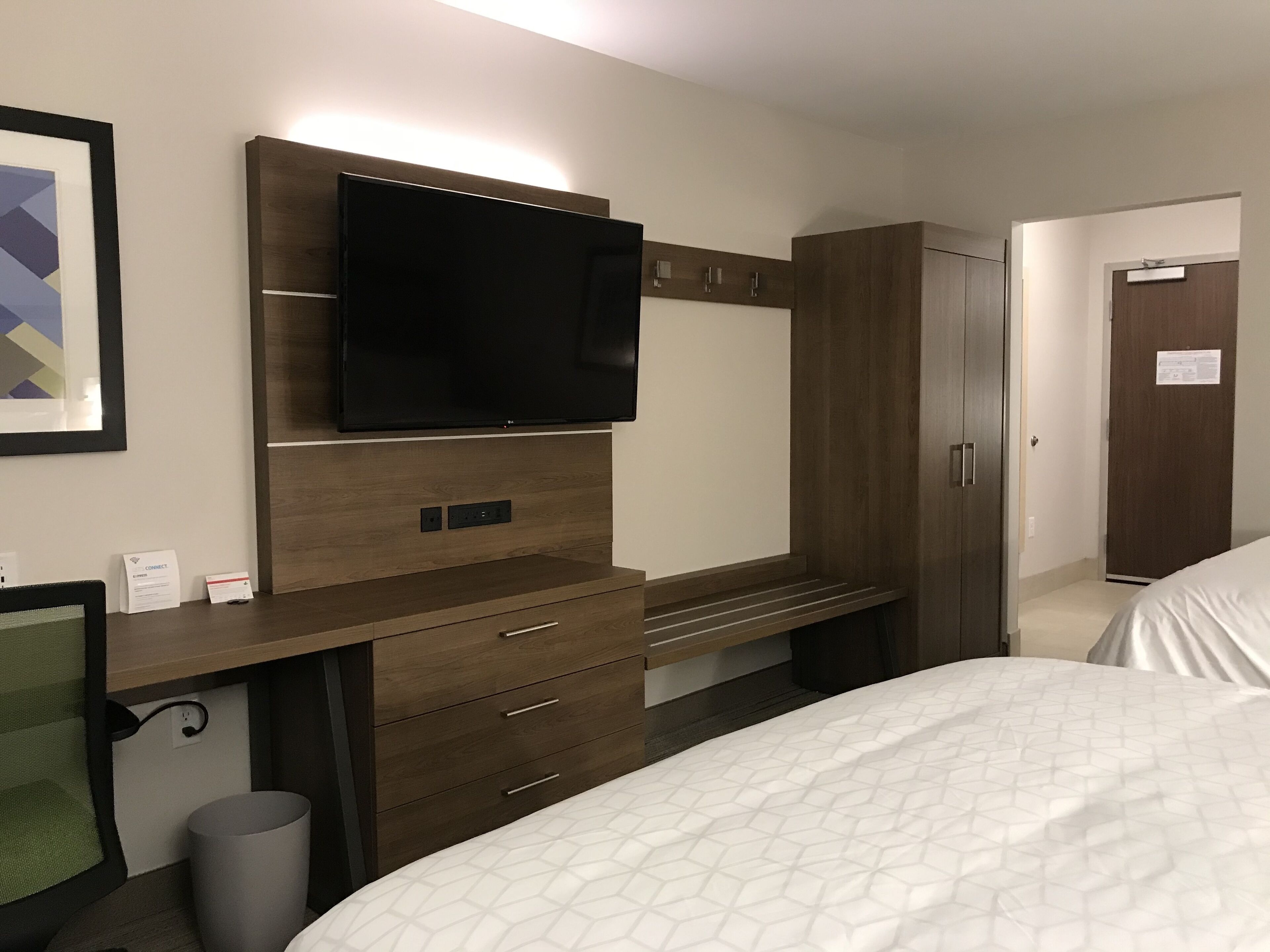 A hotel room with a large flat-screen TV mounted on the wall, a wooden dresser with drawers, a bed with a white quilt, and a door leading to another room.