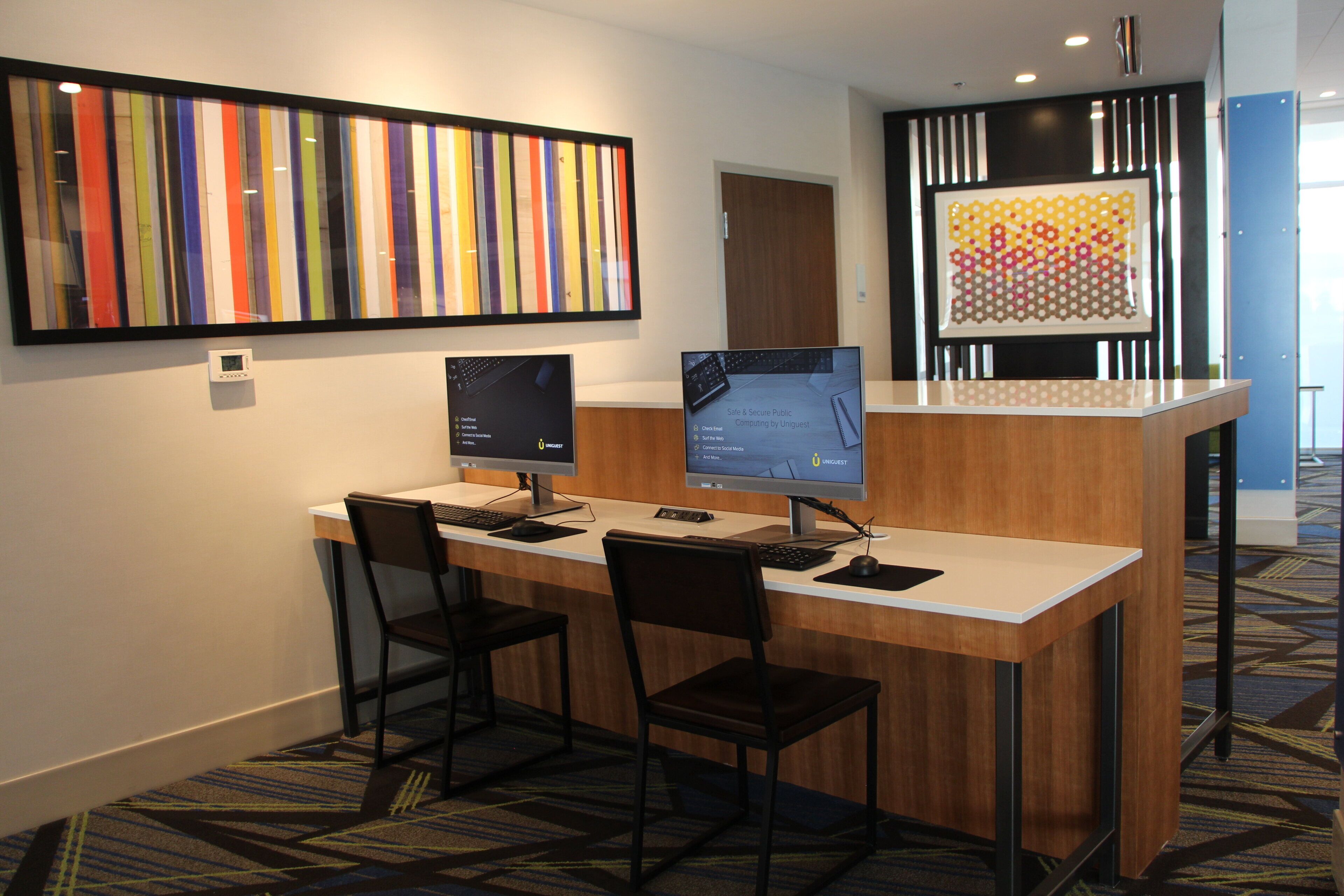 Un espace d’accueil d’hôtel comprenant deux postes de travail informatiques, une grande peinture abstraite aux couleurs vives et une moquette à motifs.
