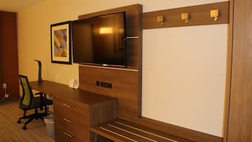 A hotel room with a wooden desk, a flat-screen TV mounted on the wall, a framed picture, and a trash bin.