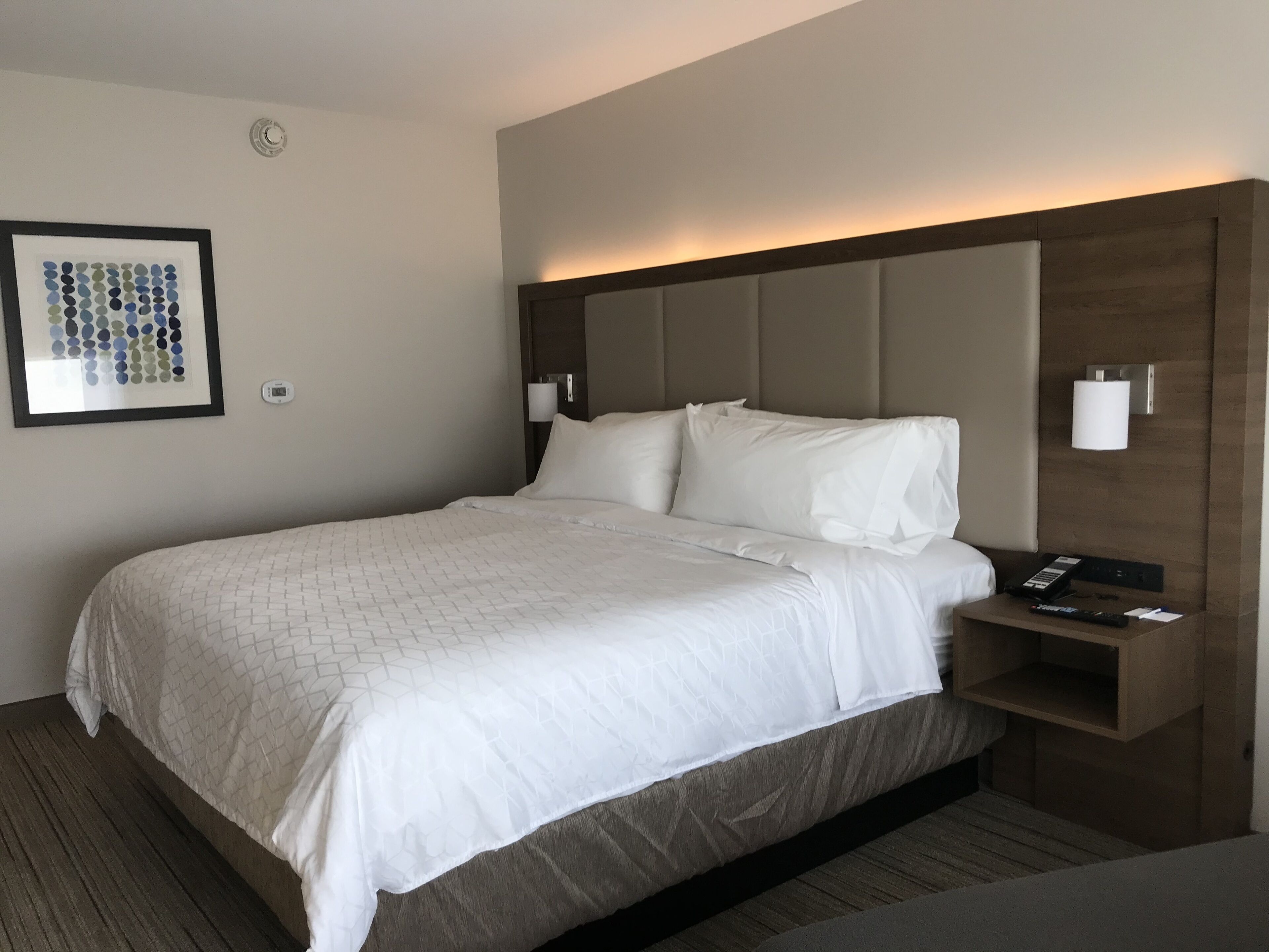 Ein Hotelzimmer mit einem großen Bett, einem Kopfteil mit integrierter Beleuchtung, einem Nachttisch mit Fernbedienung und einem gerahmten Kunstwerk an der Wand.