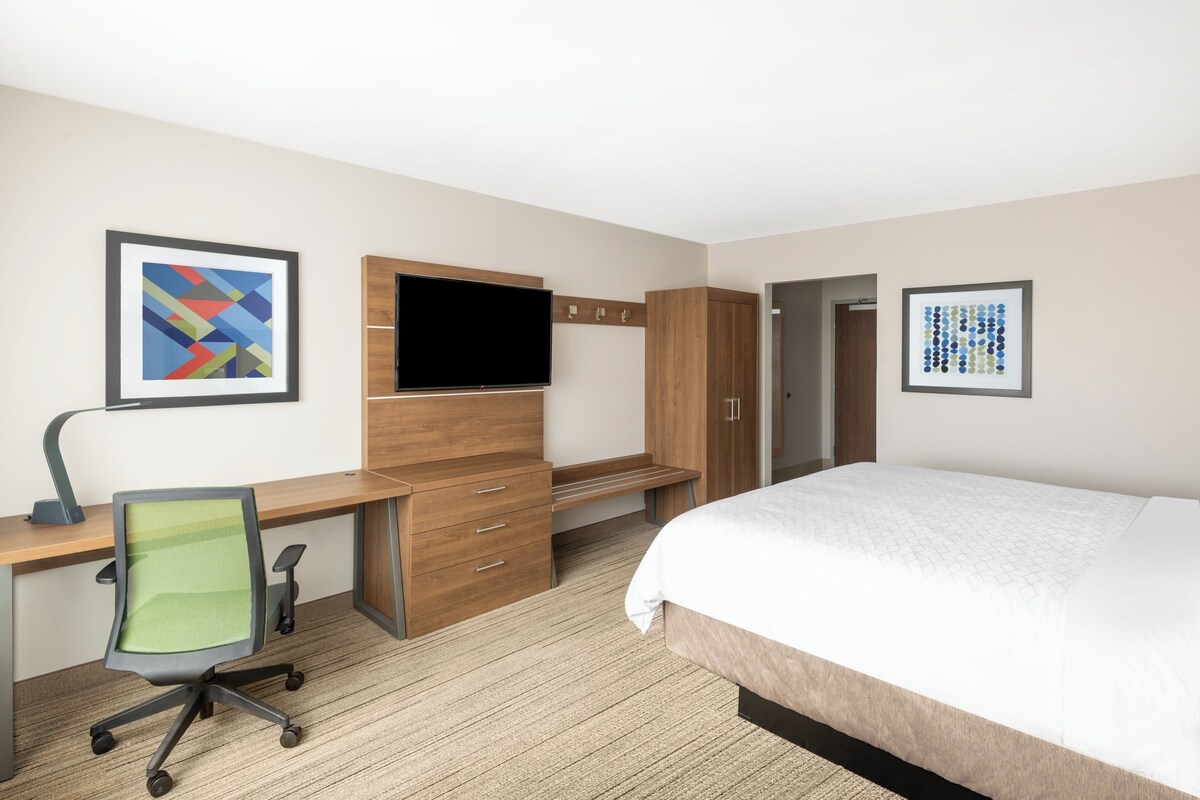 A hotel room with a bed, desk, chair, TV, wardrobe, and artwork on the walls.