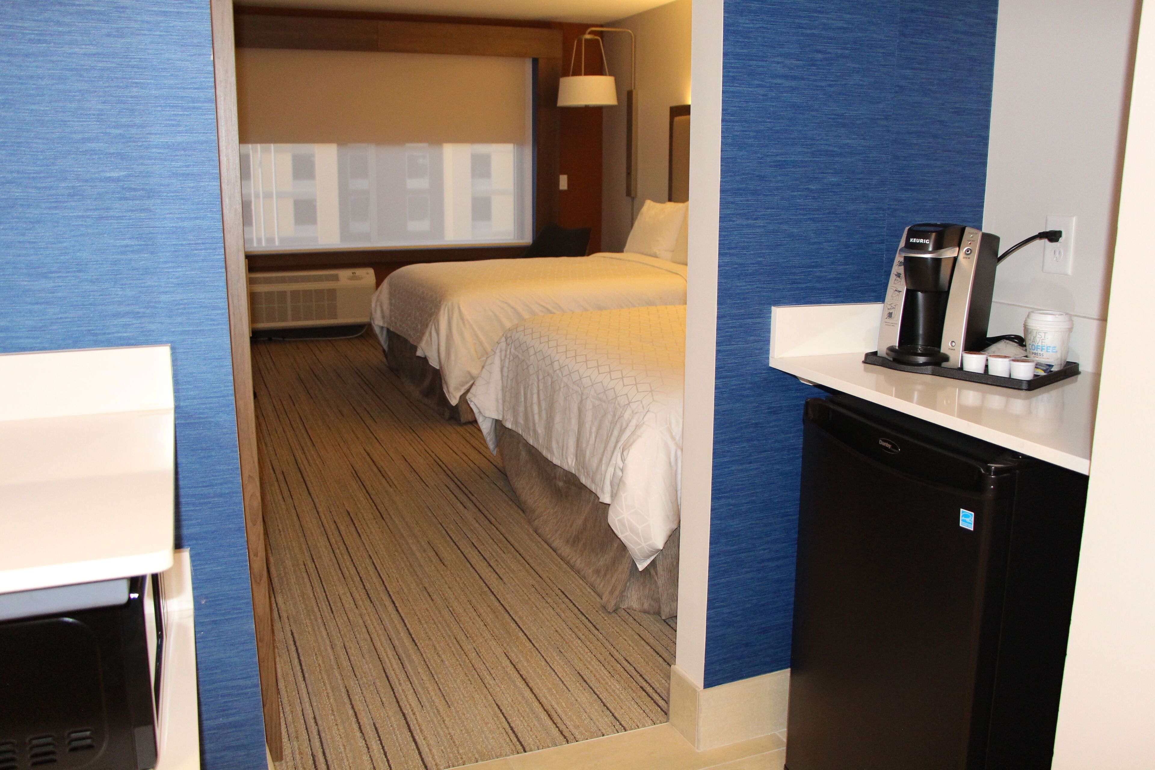 A hotel room with two beds, a small kitchenette, and a view of the cityscape.