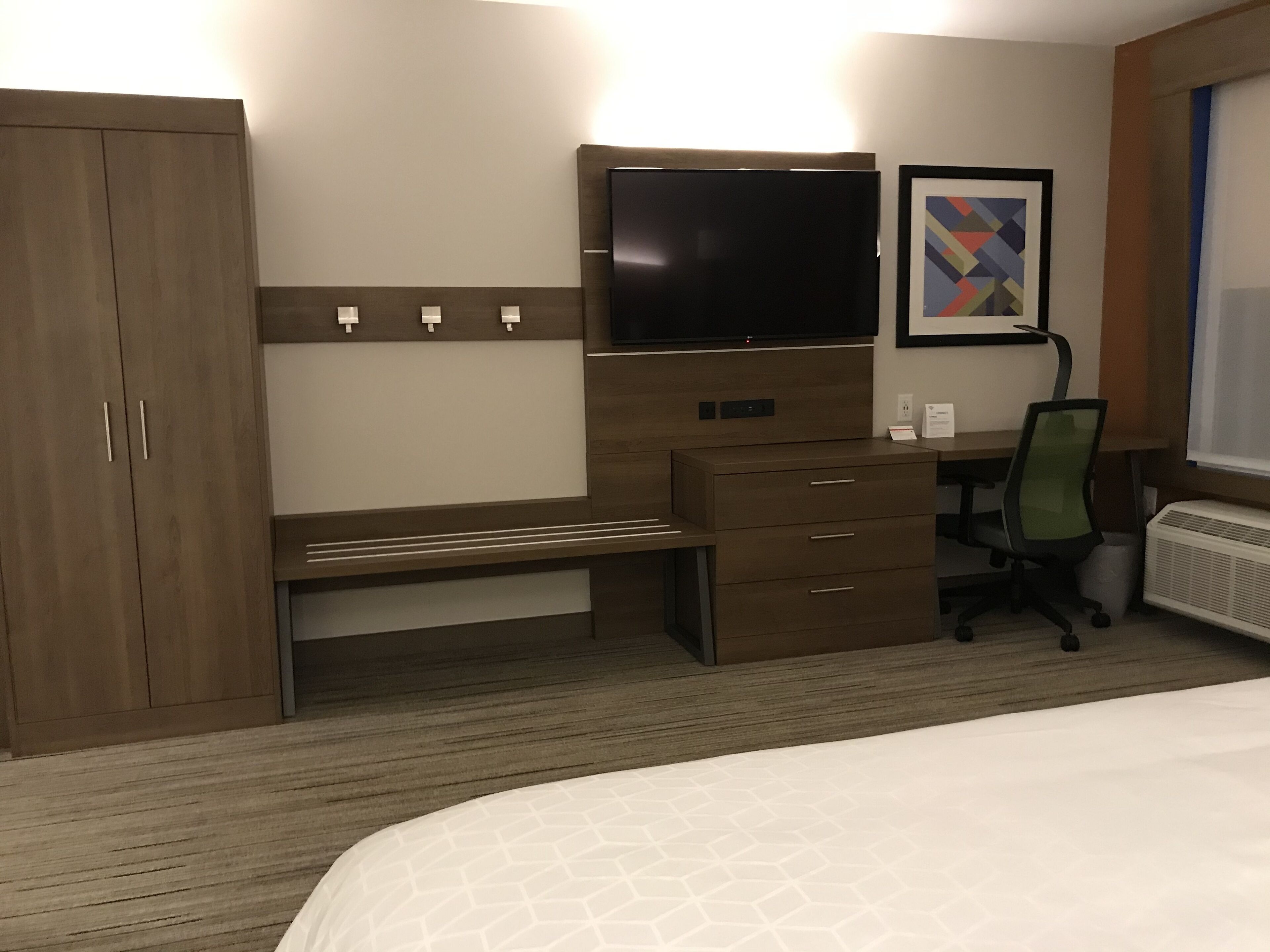 A hotel room with a bed, a desk with a chair, a wardrobe, a flat-screen TV, and a framed picture on the wall.