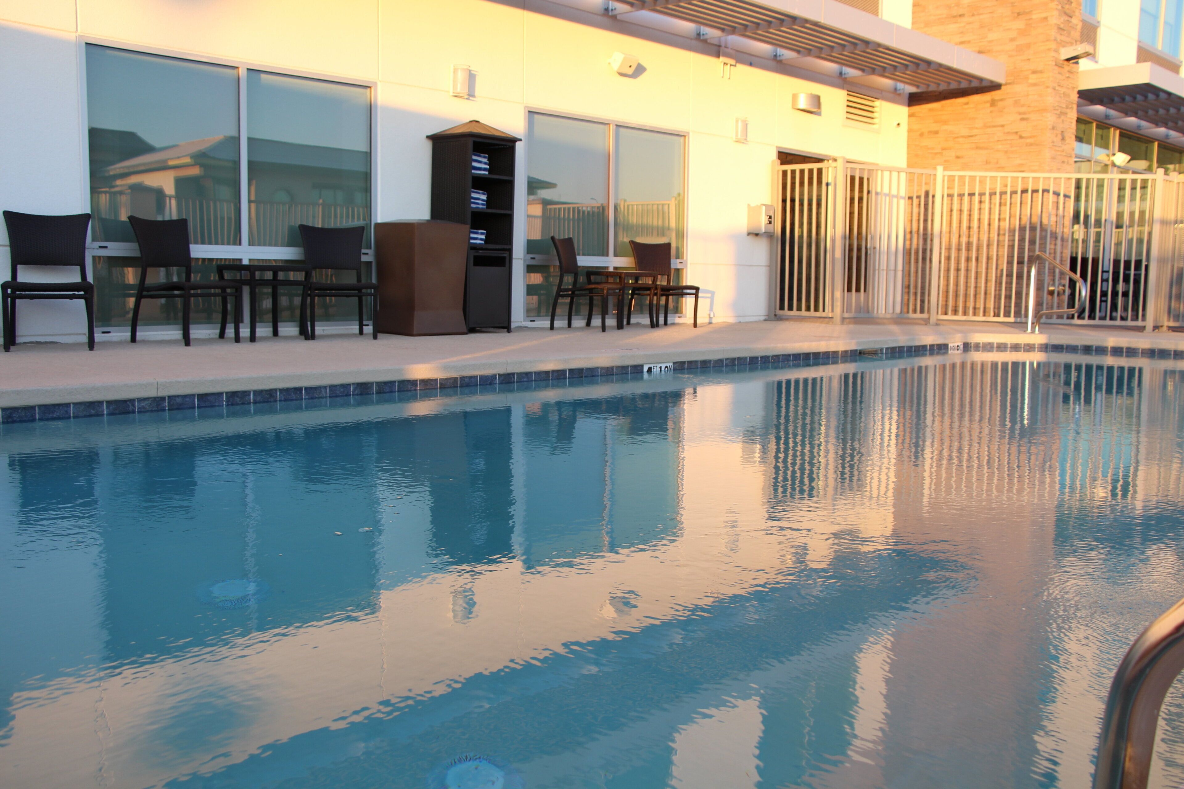 Una piscina con sillas y mesas, un ventanal de vidrio y un edificio con ventanas y un muro de ladrillo.