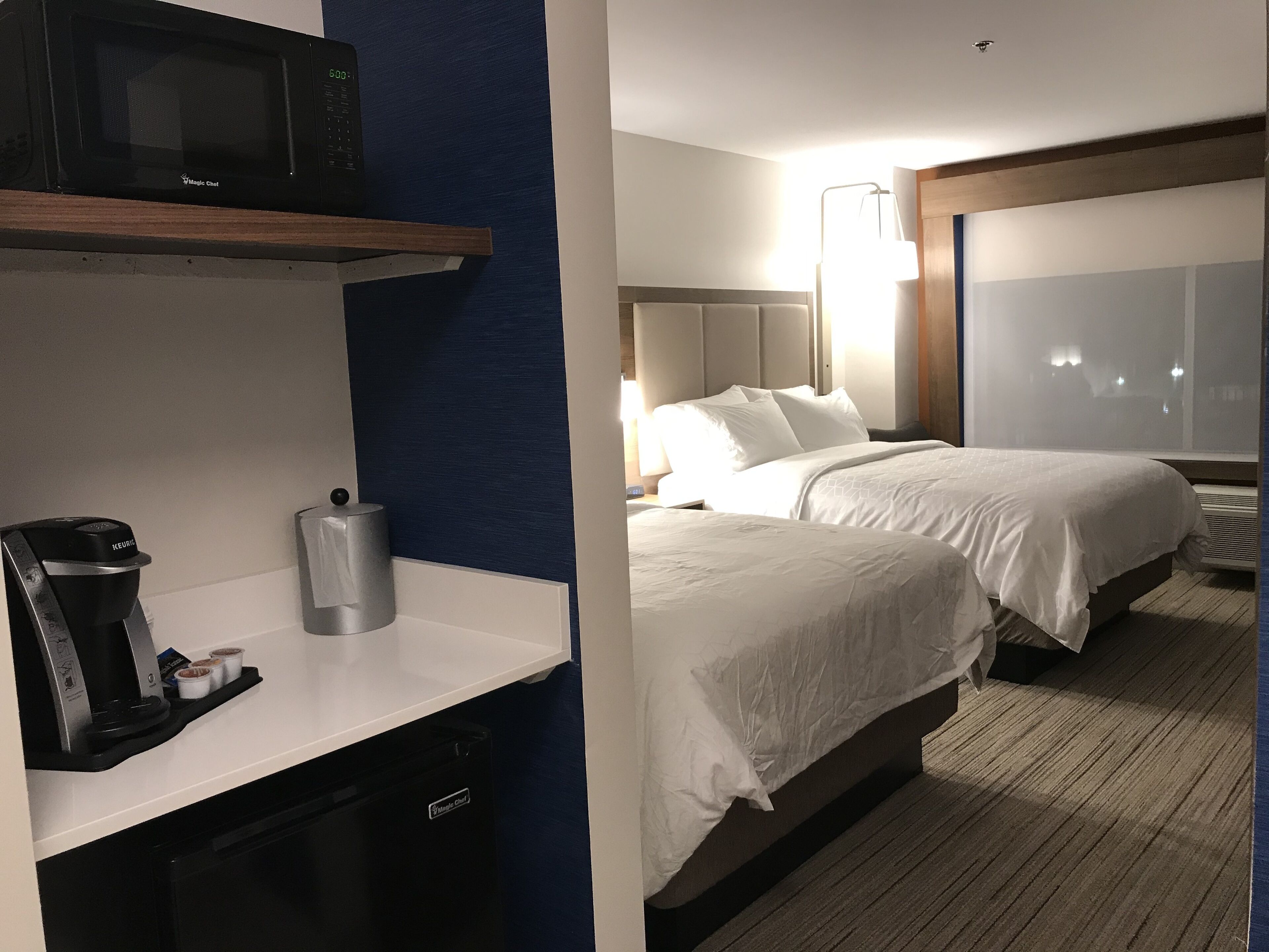 Quarto de hotel com cozinha compacta, duas camas, uma janela grande e televisão na parede.