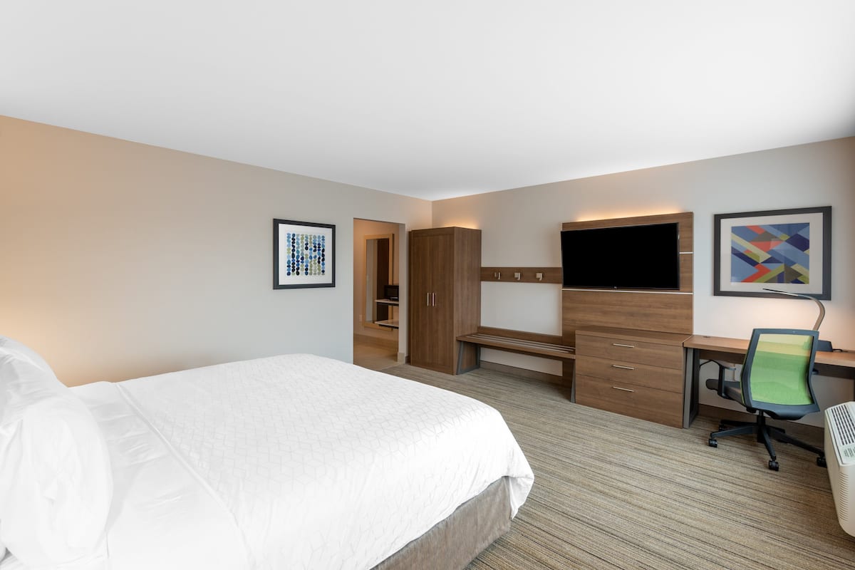 A hotel room with a large bed, a desk with a chair, a wardrobe, a television, and artwork on the walls.