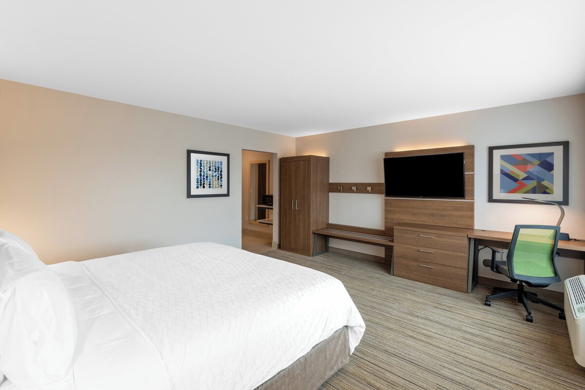 A hotel room with a large bed, a desk with a chair, a wardrobe, a television, and artwork on the walls.