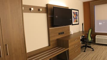 A modern hotel room with a wooden desk, a flat-screen TV mounted on the wall, a framed artwork, and a window with a view.