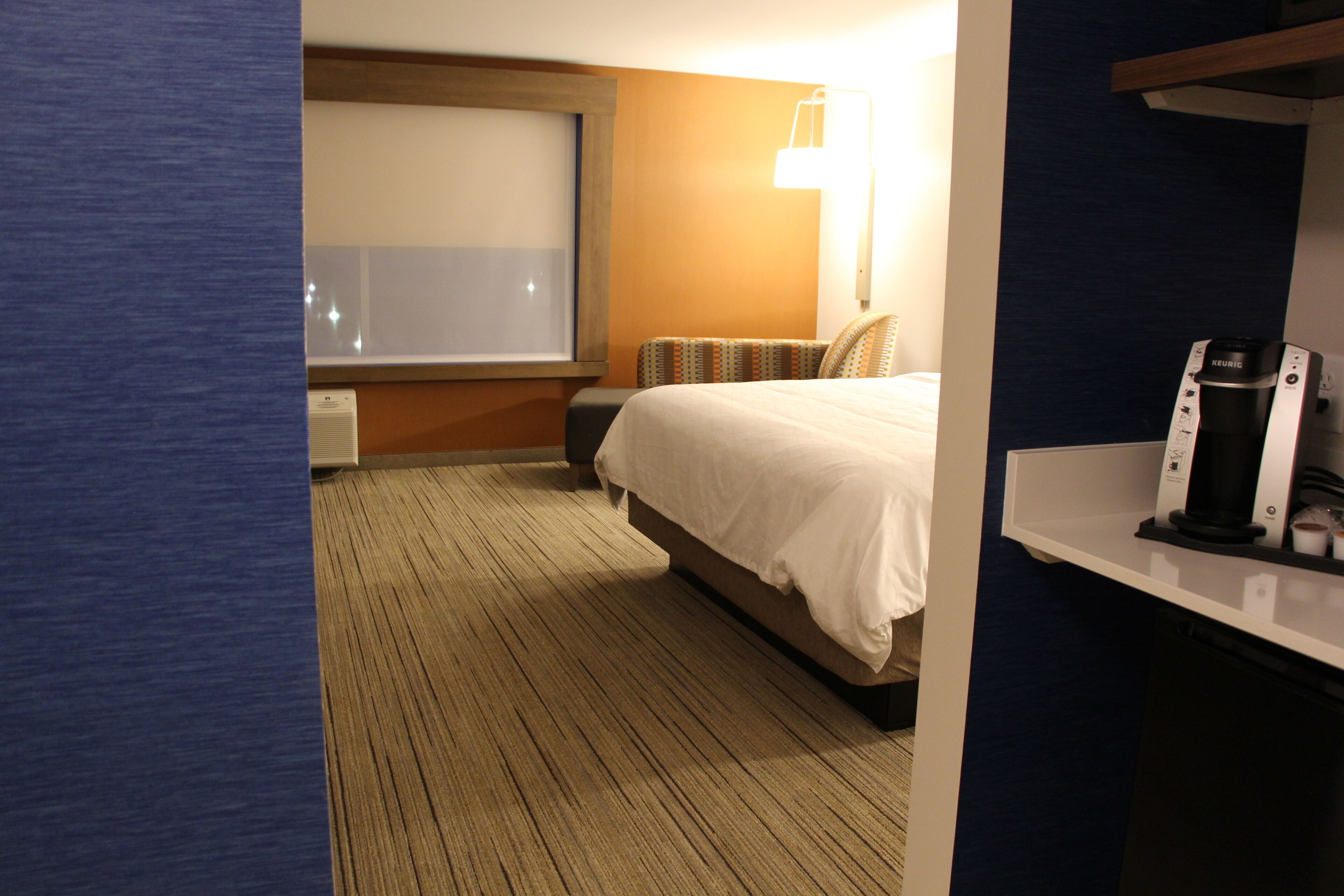 Quarto de hotel com uma cama grande, uma mesinha de cabeceira com cafeteira, uma janela e piso carpeteado.