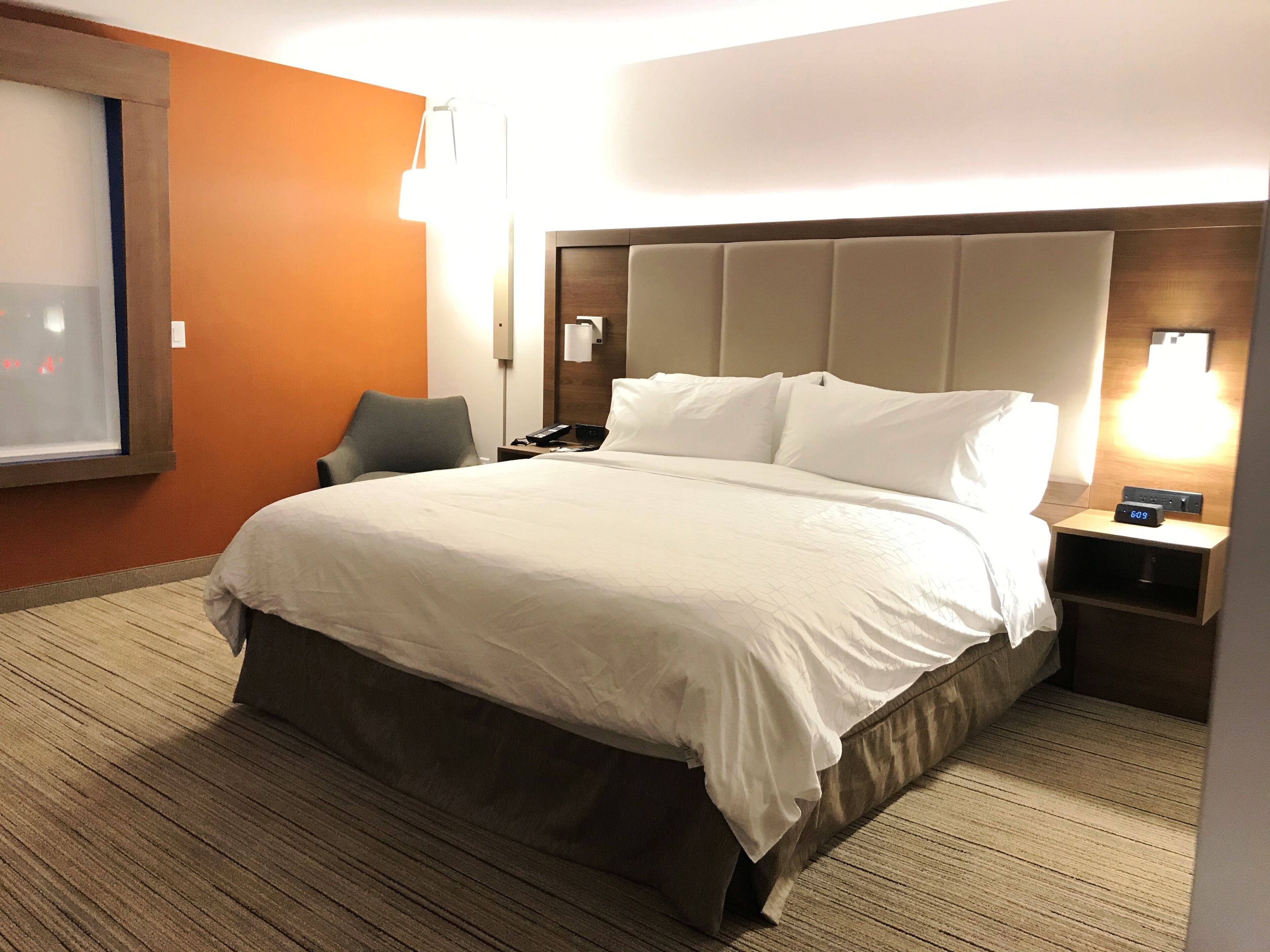 Una habitación de hotel con una cama grande, una mesita de noche con un reloj, una silla y una ventana con persianas.