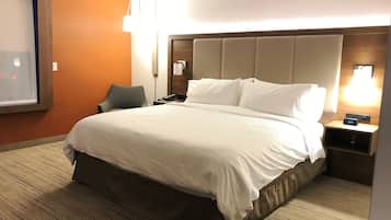 Una habitación de hotel con una cama grande, una mesita de noche con un reloj, una silla y una ventana con persianas.
