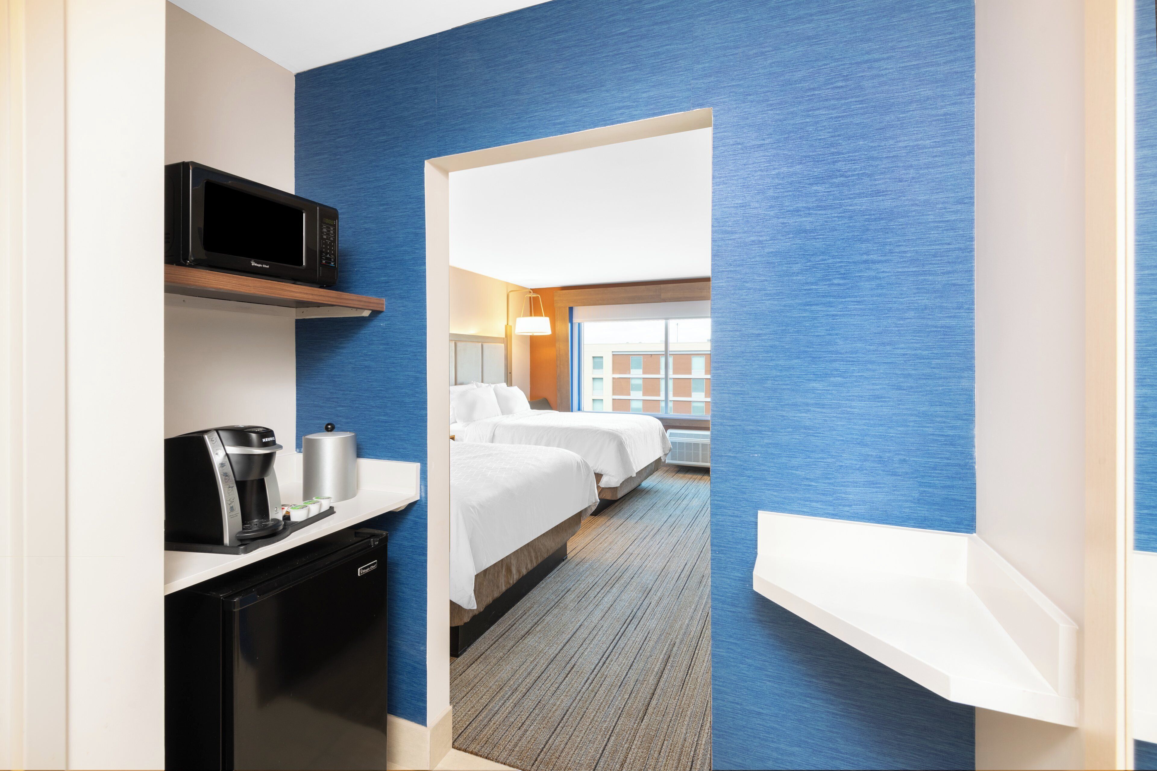 A hotel room with a kitchenette, a bed, a desk, and a view of the outside.