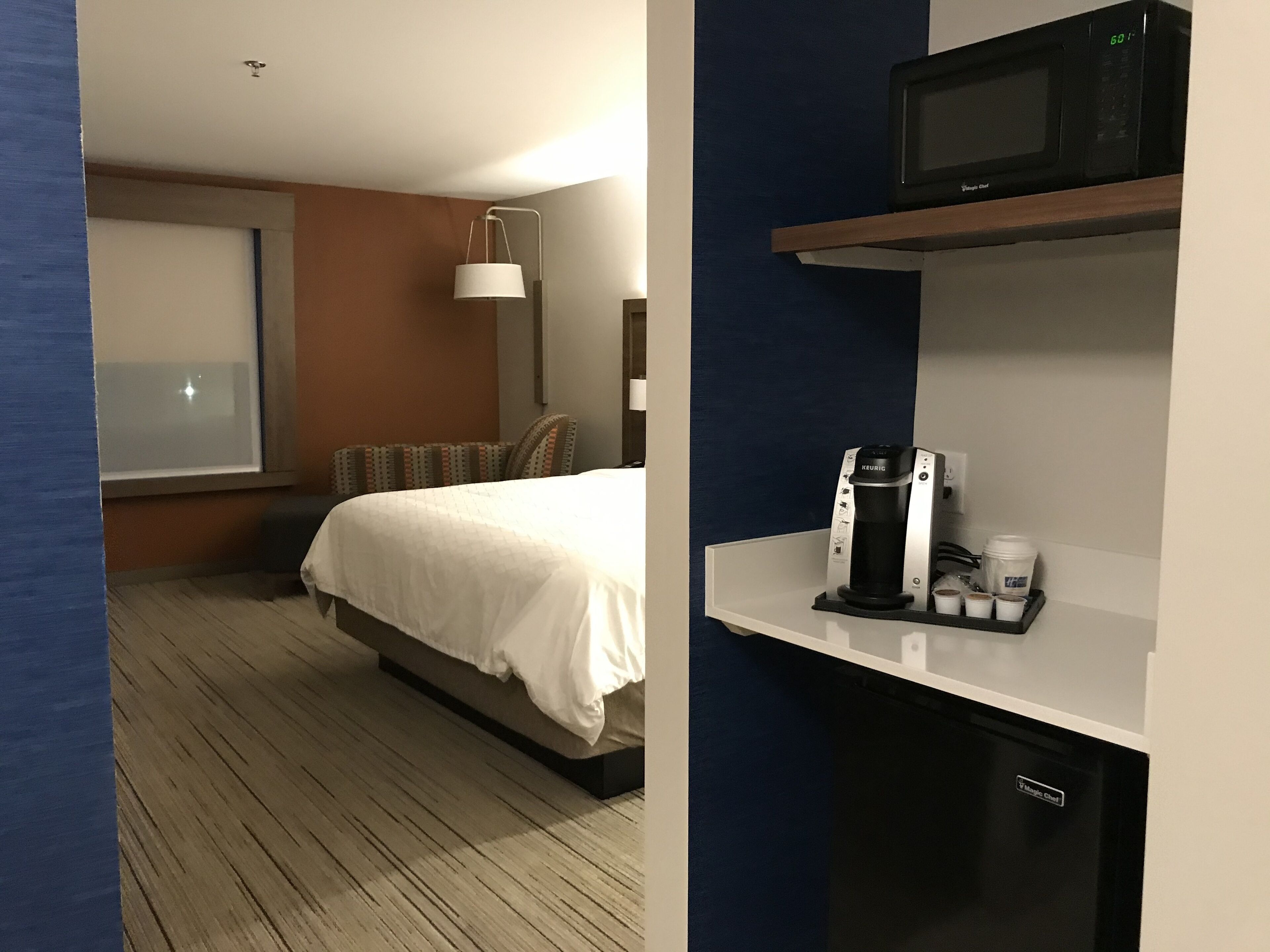 Habitación de hotel con cama, mesita de noche con cafetera, microondas y ventana con persianas.