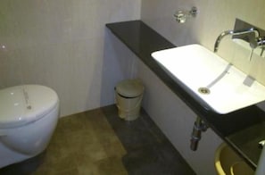 A small bathroom with a white toilet, a sink with a black countertop, and a trash bin. - Mallige Residency (Hassan)