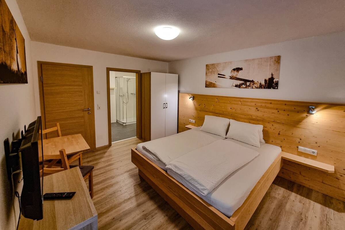 A bedroom with a wooden bed, a desk, and a chair. There is a door leading to a bathroom with a glass shower enclosure.