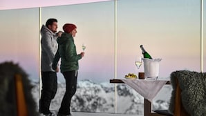A couple standing by a table with a bottle of champagne and a glass, overlooking a snowy landscape.