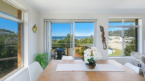 A dining area with a wooden table, white chairs, and a view of the ocean through large glass doors. - Seaview 12 - Pete & Kaz's - Modern Apartment (AVOCA BEACH)