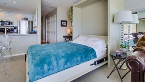 A bedroom with a bed, a desk, a chair, and a kitchenette in the background. - 3rd fl. Bayfront condo, balcony, parking, bikes (San Diego)