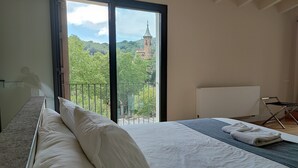 Une chambre à coucher avec un lit, une chaise, une vue sur une église à travers une porte-fenêtre donnant sur un balcon, et un tableau blanc.