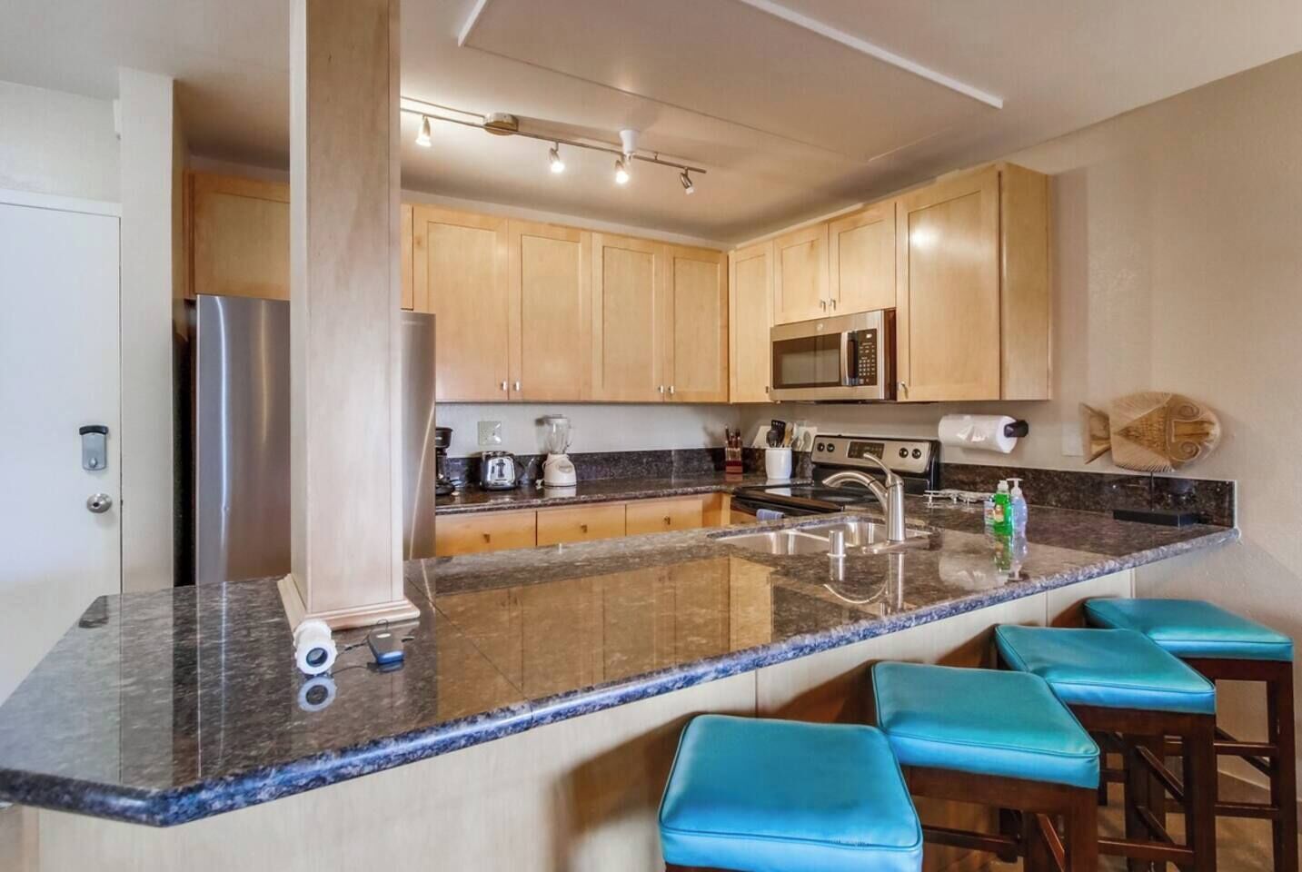 A modern kitchen with a granite countertop, stainless steel appliances, wooden cabinets, and blue bar stools.