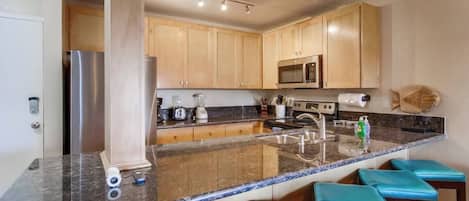 A modern kitchen with a granite countertop, stainless steel appliances, wooden cabinets, and blue bar stools.