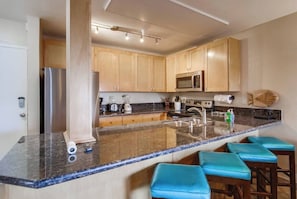 A modern kitchen with a granite countertop, stainless steel appliances, wooden cabinets, and blue bar stools. - Heart of PB steps to beach plus garage parking (San Diego)