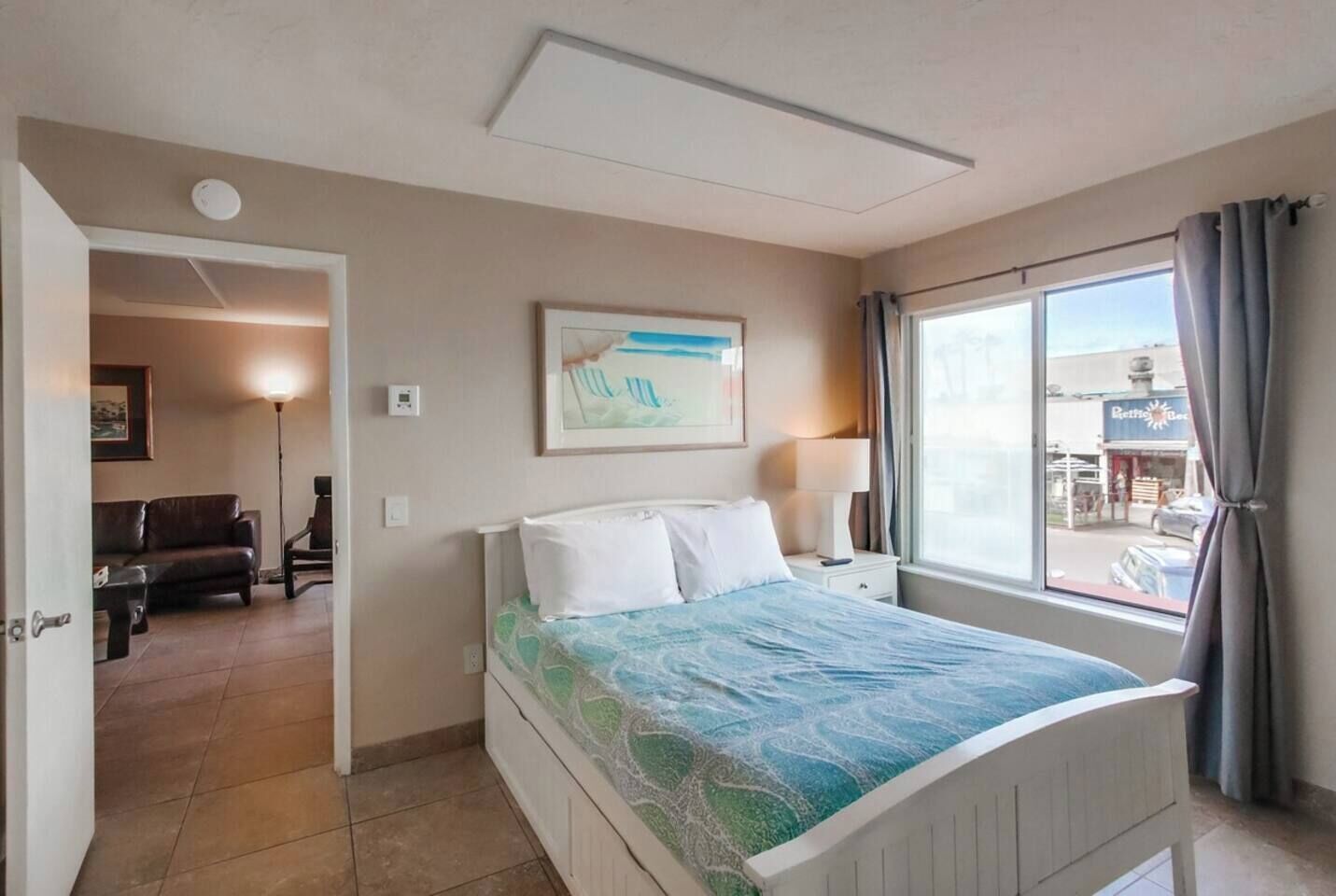 A hotel room with a bed, a nightstand, a lamp, a painting, and a view of the outside.