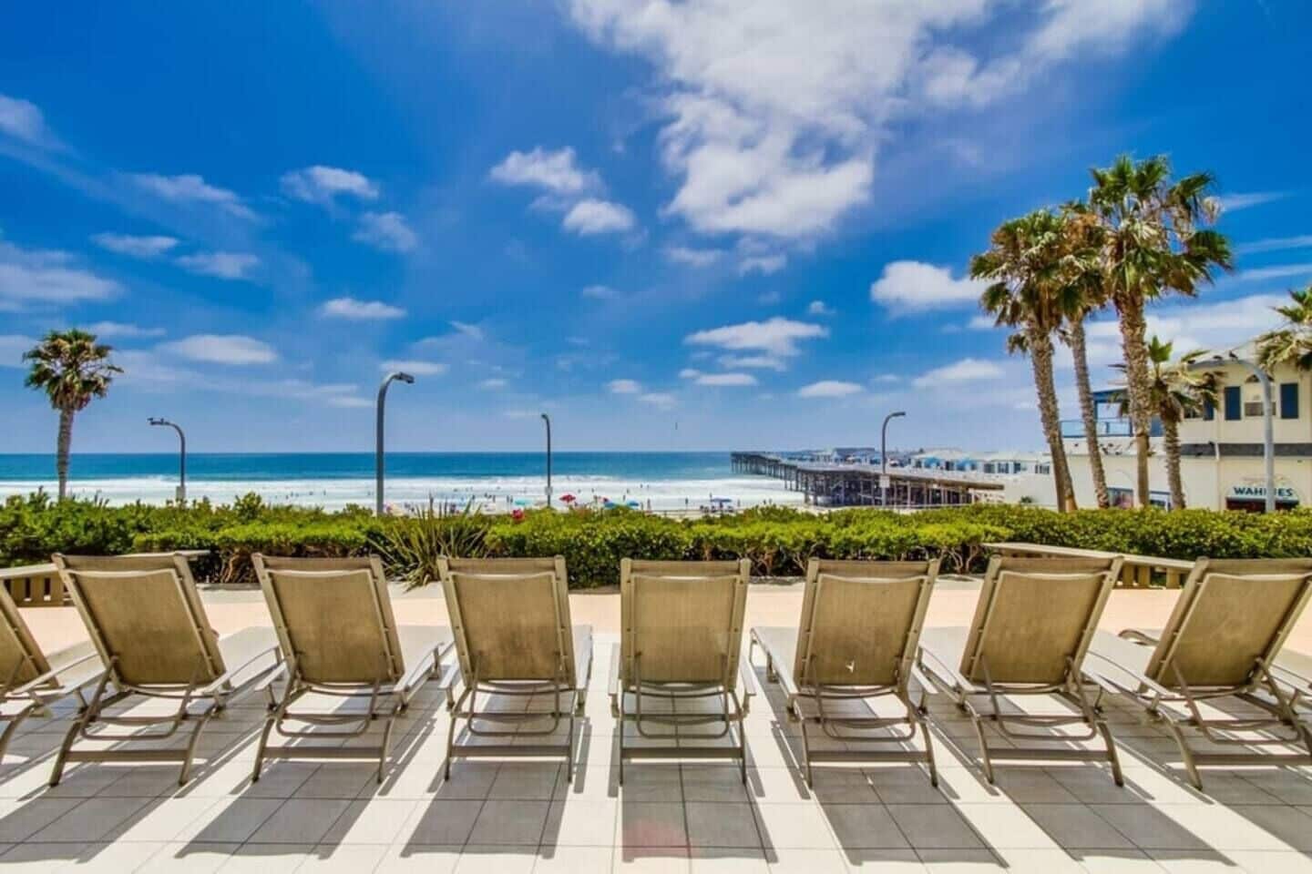 A beachside seating area with chairs facing the ocean, a pier, and palm trees.