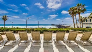 A beachside seating area with chairs facing the ocean, a pier, and palm trees.