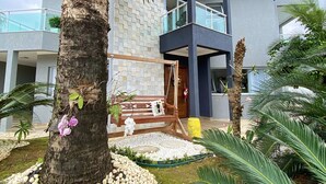 Een modern gebouw met een gevel van natuursteen, een houten bank met kussen, en een palmboom op de voorgrond.