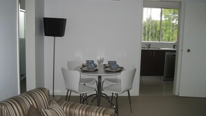 Un coin repas avec une table dressée pour deux personnes, deux chaises, une lampe sur pied et un canapé. On aperçoit une cuisine au fond.