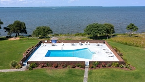 A rectangular swimming pool with a brick deck, surrounded by a grassy area and trees, with a view of the sea in the background. - Villas M1: 3 bedroom Townhome, Community Pool & Tennis Court (Nags Head)