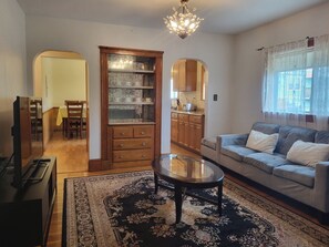 A living room with a sofa, coffee table, and a dining area with chairs and a table. There is a kitchen visible through an open doorway. - A Cozy 5BR Apartment near Harvard Square and MIT (Cambridge)