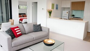 A modern living room with a grey sofa, a glass coffee table, and a kitchen area with white cabinets and a countertop.