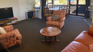 A living room with a sofa, armchair, and a round coffee table. There is a television on a stand, a small table with a lamp, and a view of the outdoors through large windows. - “CedarView at Kinloch” - a brilliant 3 bedroom holiday home with stunning views  (Kinloch)