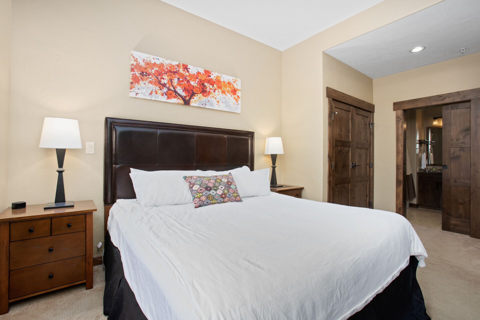 A hotel room with a large bed, two bedside tables with lamps, a wooden headboard, a colorful wall art piece, and a wooden door.