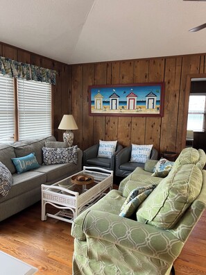 A living room with a wooden paneled wall, a painting of beach huts, and a grey sofa with patterned cushions.