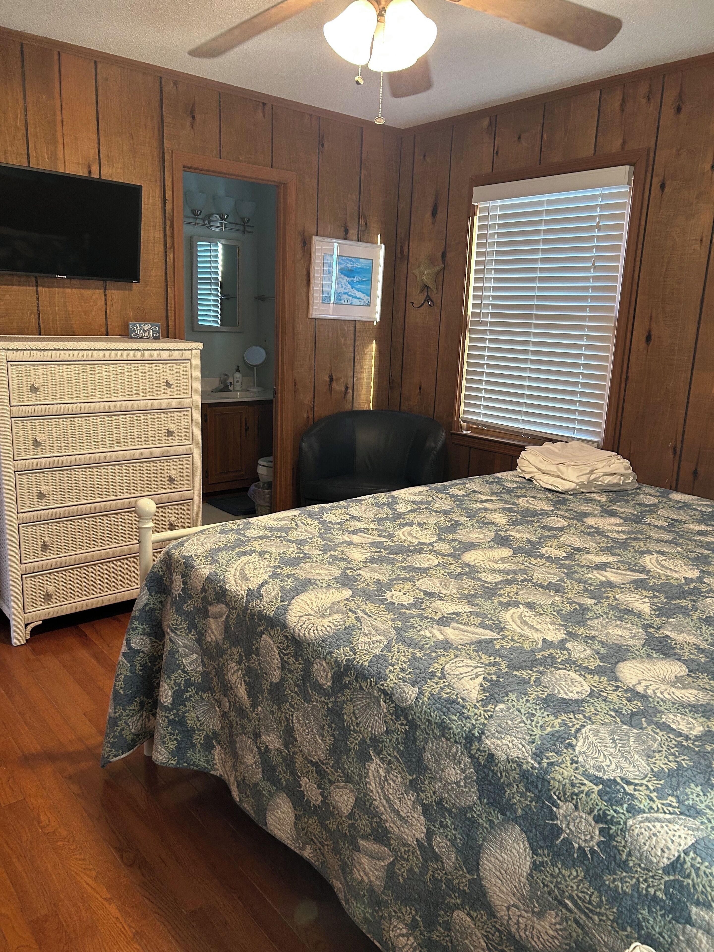A bedroom with wooden paneling, a bed with a patterned bedspread, a dresser, a chair, a ceiling fan, and a window with blinds.