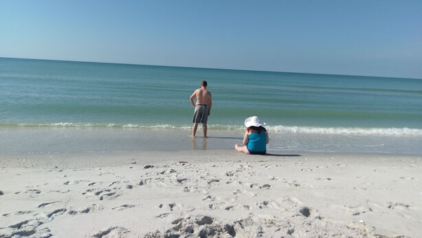 Ein Strand mit einer stehenden und einer sitzenden Person im Sand, beide dem Meer zugewandt.