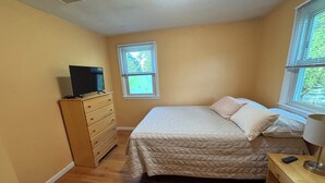A bedroom with a bed, dresser, television, windows, and a lamp.