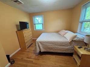 A bedroom with a bed, dresser, television, windows, and a lamp.