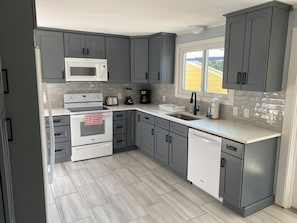 Uma cozinha moderna com armários cinza, micro-ondas branco, fogão, lava-louças e uma janela com vista.