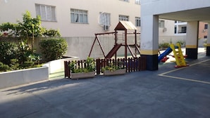 Ein Spielplatz mit Schaukel und Rutsche, umgeben von einem niedrigen Zaun und Kübelpflanzen.
