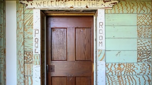 A wooden door with a 'WELCOME TO THE ROOM' sign above it, surrounded by patterned wallpaper.