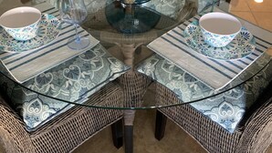 Une table ronde en verre, surmontée d’un vase bleu au centre, entourée de quatre chaises en osier dotées de coussins à motifs.