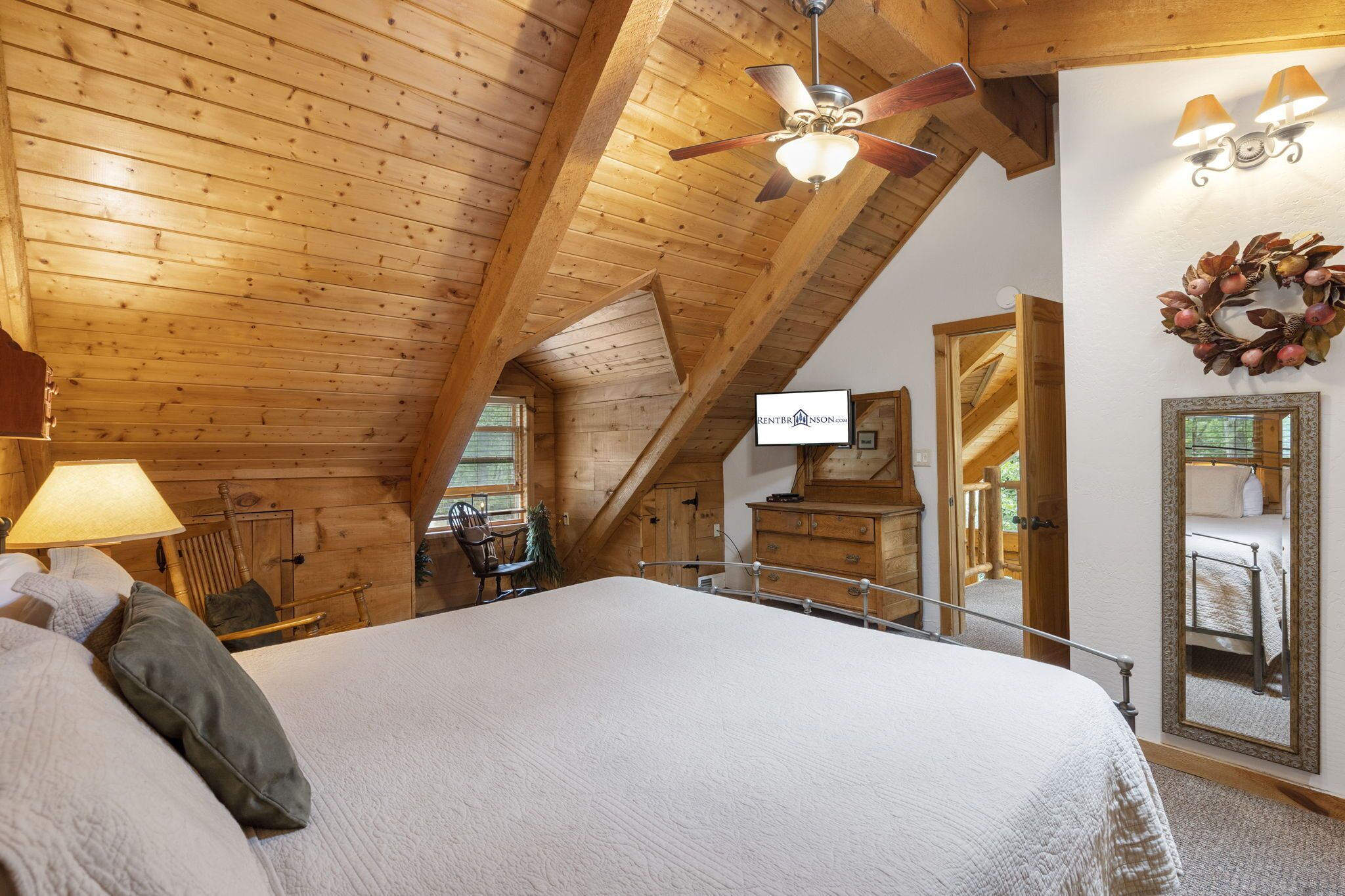 Een gezellig slaapkamer onder de dakpannen met een groot bed, houten balken, een plafondventilator, een spiegel en uitzicht op buiten.