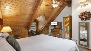 A cozy attic bedroom with a large bed, wooden ceiling beams, a ceiling fan, a mirror, and a view of the outside.